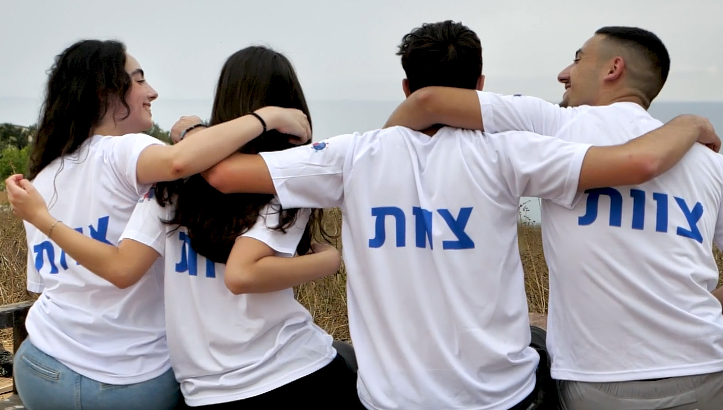 Four teenagers in a line facing away from the camera with their arms wrapped around each other and looking into distance or at each other.
