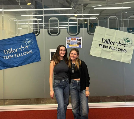 Two alumni standing in front of a glass wall with Diller Teen Fellows flags behind them.