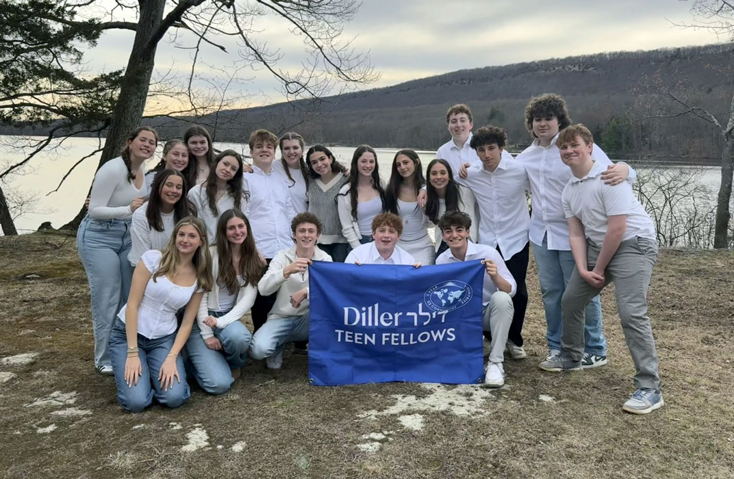 Group of teens in white shirts standing on a river bank holding a blue flag that reads Diller Teen Fellow