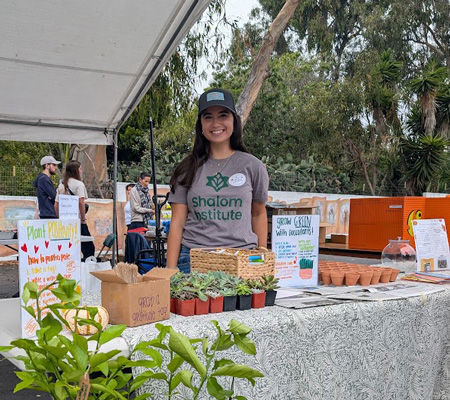 Woman wearing a Shalom Institute t-shirt at an outdoor booth with succulents, signs, and planting supplies.