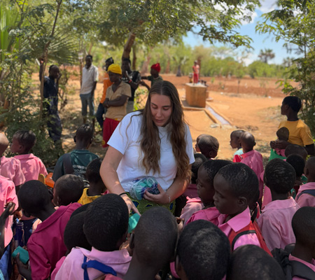 Woman distributes items to children in pink uniforms near trees and water pump in rural setting.