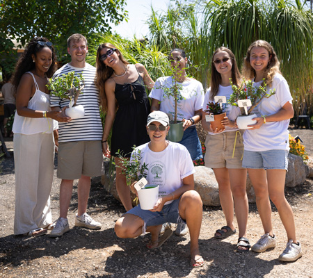 Seven people hold potted plants on dirt path in garden area with rocks and greenery